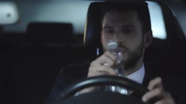 takım elbiseli mutlu sakallı adam seçici odak araba sürüş, şişe alma, su içme ve saat bakarak 