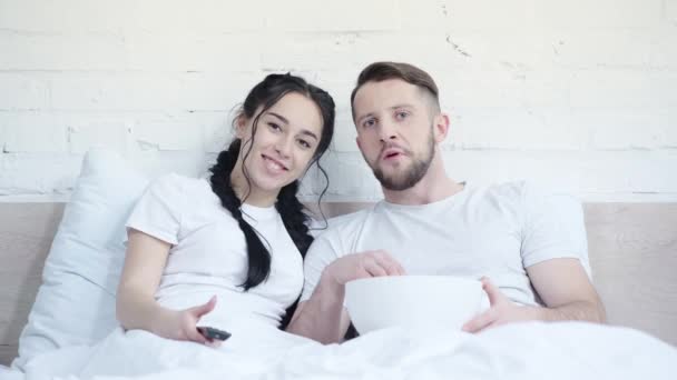 有吸引力的和快乐的女人看电影附近的英俊的男朋友和吃爆米花在卧室 — 图库视频影像