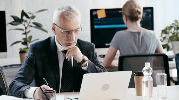 选择性的焦点英俊的胡子男子在眼镜写作 而使用笔记本电脑在办公室附近的女人 — 图库视频影像