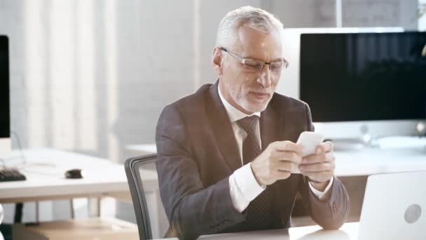メガネでハンサムなあごひげの男は笑顔で オフィスでスマートフォンを使用しています — ストック動画