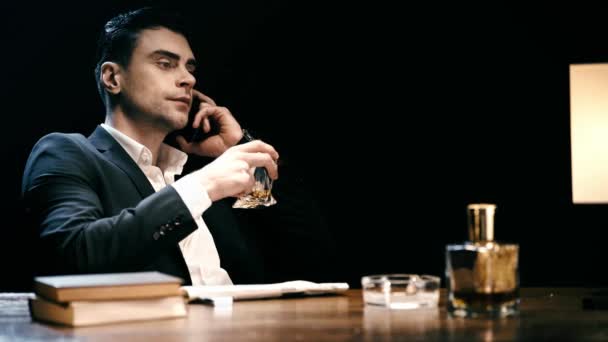 テーブルに座り スマートフォンで話し 黒で孤立したウイスキーを飲む真面目なビジネスマン — ストック動画