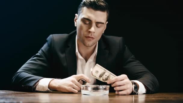 黒で隔離された木製のテーブルで灰皿の上にライターと燃えるドル紙幣を保持しているビジネスマン — ストック動画