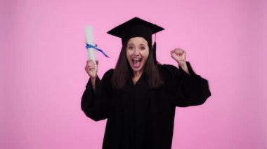 Akademik cüppe mutlu öğrenci eliyle tezahürat ve kamerada diploma gösteren pembe üzerinde Izole
