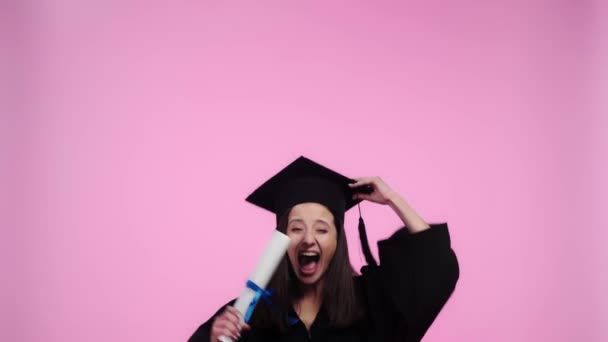 兴奋的学生在学术礼服看着相机和跳跃与毕业帽和文凭孤立在粉红色 — 图库视频影像