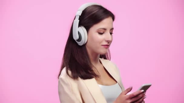 美丽的微笑的女孩在耳机与智能手机听音乐和跳舞孤立在粉红色 — 图库视频影像
