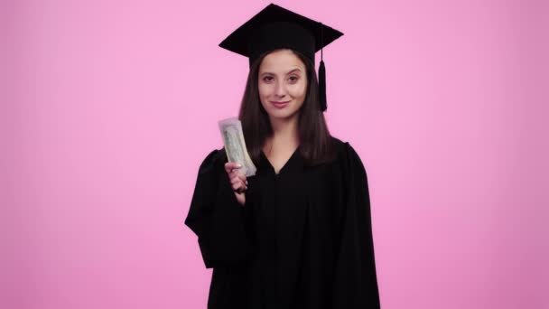 beautiful student in academic gown looking at camera, smiling, giving money to woman, taking diploma and winking isolated on pink
