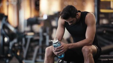 Sakallı sporcu spor salonunda müzik dinliyor ve su içiyor. 