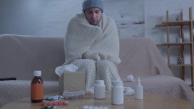 Hastalıklı bir adam hapşırırken masanın yanında ilaçlarla oturuyor. 