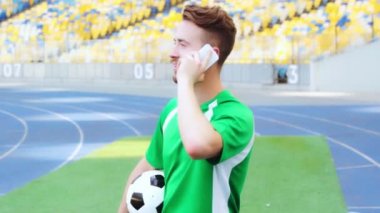 Gülen futbolcu topu tutuyor ve akıllı telefondan konuşuyor. 