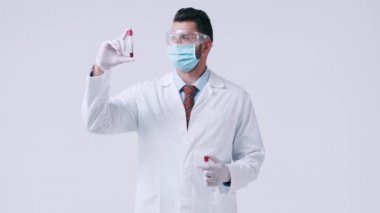 Tıbbi maskeli doktor beyaz üzerinde kan olan test tüplerine bakıyor.