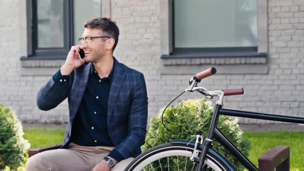 眼鏡をかけた幸せな男が手を振って自転車の近くのスマートフォンで話しています — ストック動画