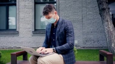 Maskeli serbest çalışan, bankta otururken dizüstü bilgisayar kullanıyor. 