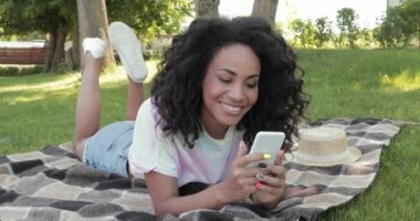 Gülümseyen Afrikalı Amerikalı kız parkta ekose üzerinde akıllı telefon kullanıyor. 