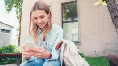 Kentteki bankta akıllı telefon kullanan gülümseyen öğrenci 