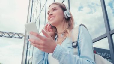 Kulaklıklı neşeli bir öğrenci elinde akıllı telefon ile şehir caddesindeki binanın yanında.
