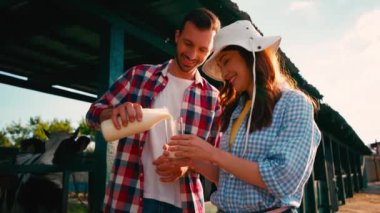 Erkek, ineğin yanındaki şapkalı kadının bardağına taze süt döker.
