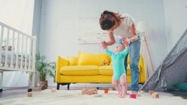 Anne el ele tutuşuyor ve bebeğe oyuncakların yanında battaniyede yürümesinde yardım ediyor.