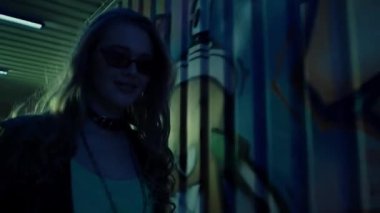 Güneş gözlüklü genç bir kadın duvarın yanında grafiti ile yürüyor. 