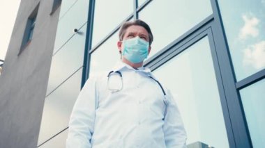 Sağlık maskeli doktorun düşük açılı görüntüsü dışarıda kliniğin yanında duruyor.