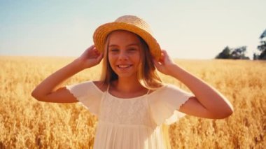 Beyaz elbiseli kız hasır şapkaya dokunuyor ve buğday tarlasında gülüyor. 