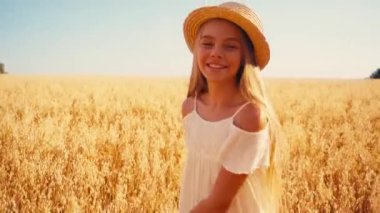Beyaz elbiseli kız dönüp buğday tarlasında hasır şapkaya dokunuyor. 