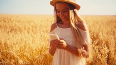 Beyaz elbiseli ve hasır şapkalı neşeli kız buğday tarlasında akıllı telefon kullanıyor. 