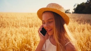 Beyaz elbiseli ve hasır şapkalı çocuk buğday tarlasında akıllı telefondan konuşuyor.