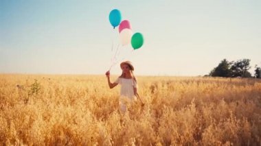 Beyaz elbiseli ve hasır şapkalı çocuk buğday tarlasında renkli balonlarla yürüyor.