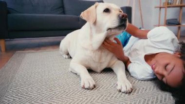 Afro-Amerikalı bir kız evdeki halıda yalan söylüyor ve köpeği okşuyor.