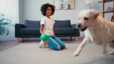Afro-Amerikalı kız evde halıda otururken top köpeğe atıyor.