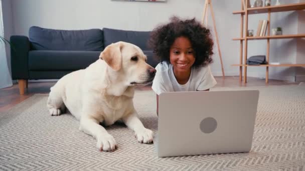 自宅でカーペットの上に横たわっている間 ラップトップを見て犬の近くにアフリカ系アメリカ人の女の子 — ストック動画