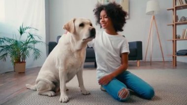 Afro-Amerikalı kız köpeğini okşuyor ve halıda otururken kameraya el sallıyor.