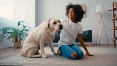 Gülümseyen Afrikalı Amerikalı kız evde halıda otururken köpeğini okşuyor.