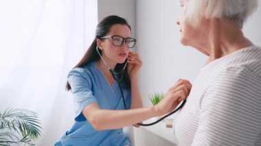 Yaşlı bir kadını steteskopla muayene eden genç bir yaşlı doktor.