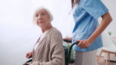 Yaşlı bir hemşire tekerlekli sandalyedeki yaşlı bir kadını taşıyor. 