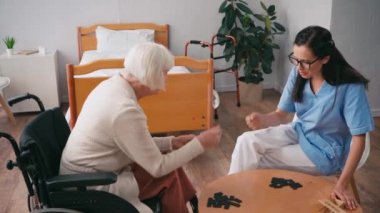 Tekerlekli sandalyedeki kadın ve hemşire dominoların yanında taş-kağıt-makas oynuyorlar.