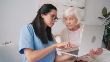 Esmer yaşlı hemşire yaşlı kadına dizüstü bilgisayarın nasıl kullanılacağını gösteriyor.