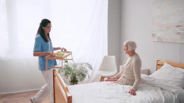 若い看護師がベッドで女性に朝食を持ってトレイを運ぶ — ストック動画