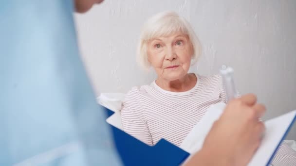 医師がクリップボードに書き込み背景がぼやけている年老いた女性と話すと — ストック動画