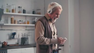 Evde akıllı telefonlu dans eden kulaklıklı mutlu bir emekli.