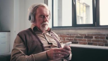 Beyaz saçlı, kulaklıklı ve akıllı telefonlu bir adam evde müzik dinliyor.
