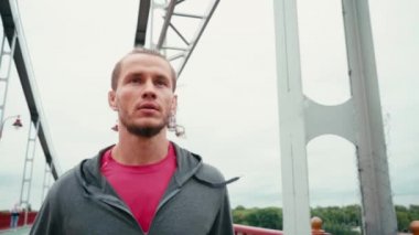 Spor kıyafetlerine odaklanmış bir adam köprüde koşuyor ve arka planda bulutlu bir gökyüzü var.
