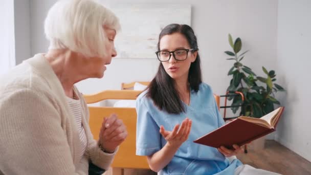 病院で本を議論する若い看護師と高齢者の女性 — ストック動画