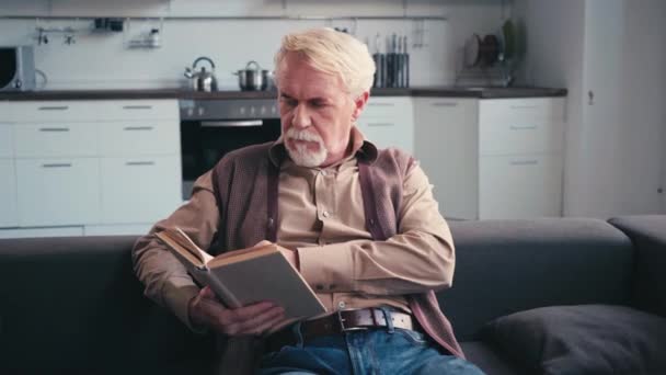 Šedovlasý Muž Čtení Knihy Zatímco Sedí Pohovce Kuchyní Pozadí — Stock video