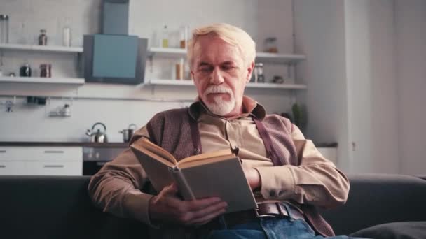 Hombre Mayor Leyendo Libro Mientras Está Sentado Sofá Con Cocina — Vídeos de Stock