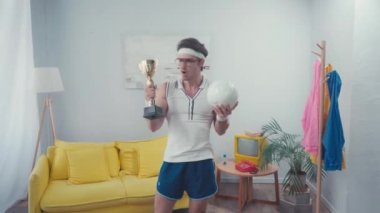 Sporcu elinde kupa ve spor topu tutarken, evde dikiliyor.