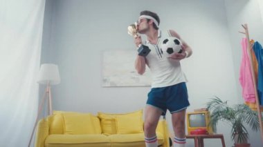 Oturma odasında spor topu ve kupayla dans eden enerjik futbolcu.