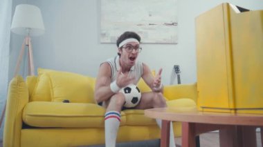 Futbol fanatiği, futbol topu takımda tezahürat yaparken, kanepede oturuyor.