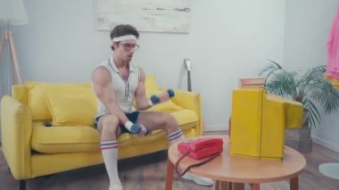 Spor adamı oturma odasında televizyon izlerken dambılları kaldırıyor.