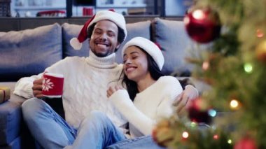 Afrika kökenli Amerikalı bir adam elinde fincanla bir kadınla Noel ağacının yanında gülüyor.